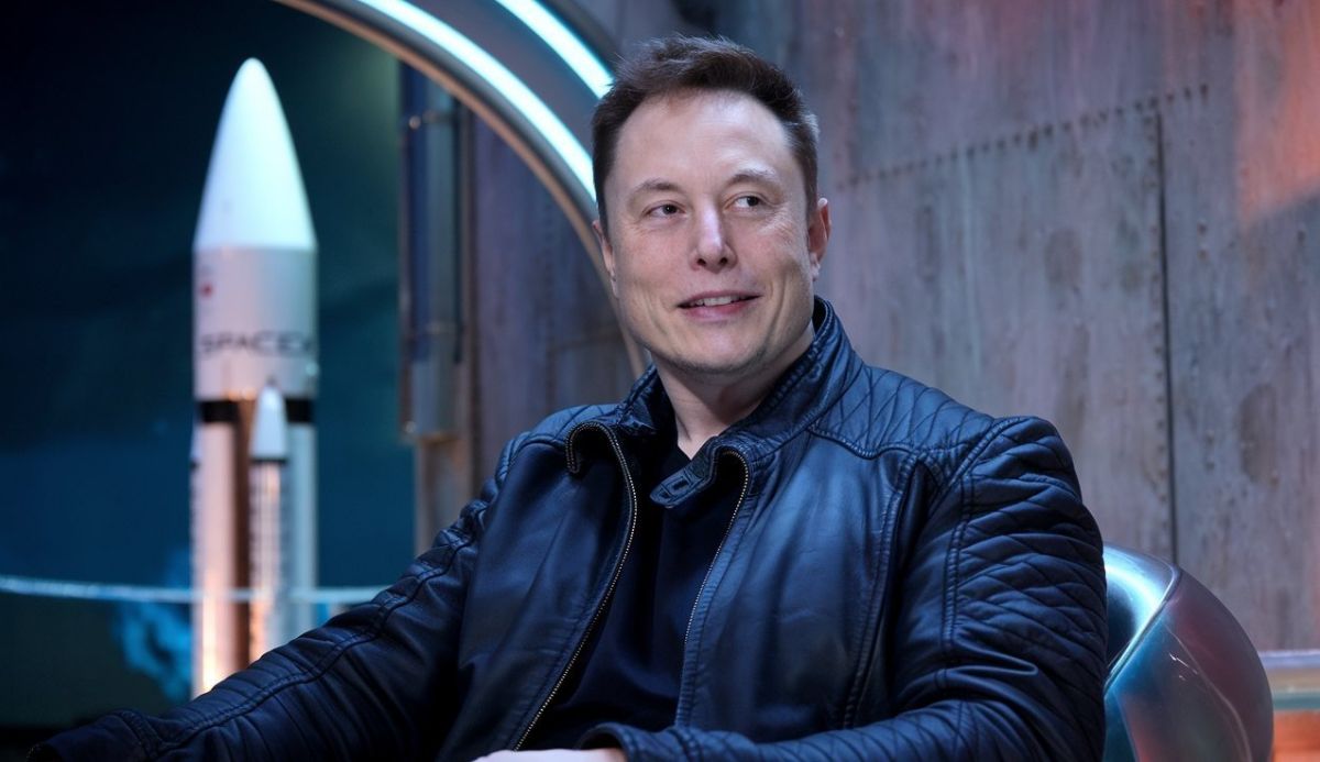 Elon Musk sitting in front of a SpaceX rocket model, wearing a black leather jacket, with a slight smile.