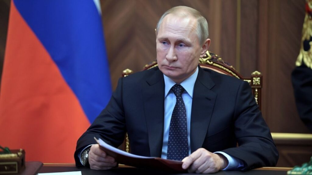 Vladimir Putin sitting at a desk with a document in hand, in front of the Russian flag, during an official meeting.