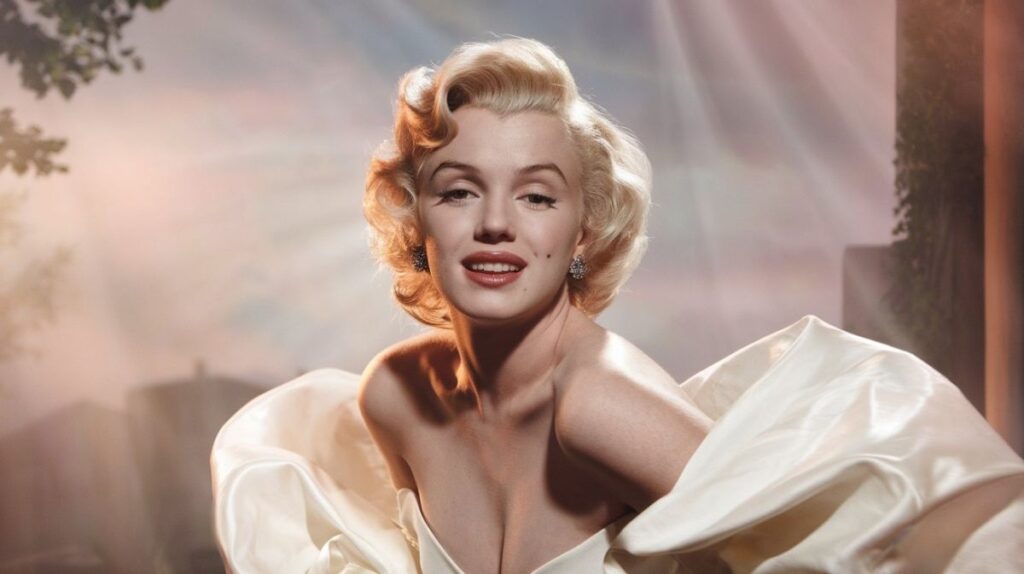 A medium-shot portrait of Marilyn Monroe, wearing a white gown, with blonde, curly hair and glamorous makeup, smiling warmly.